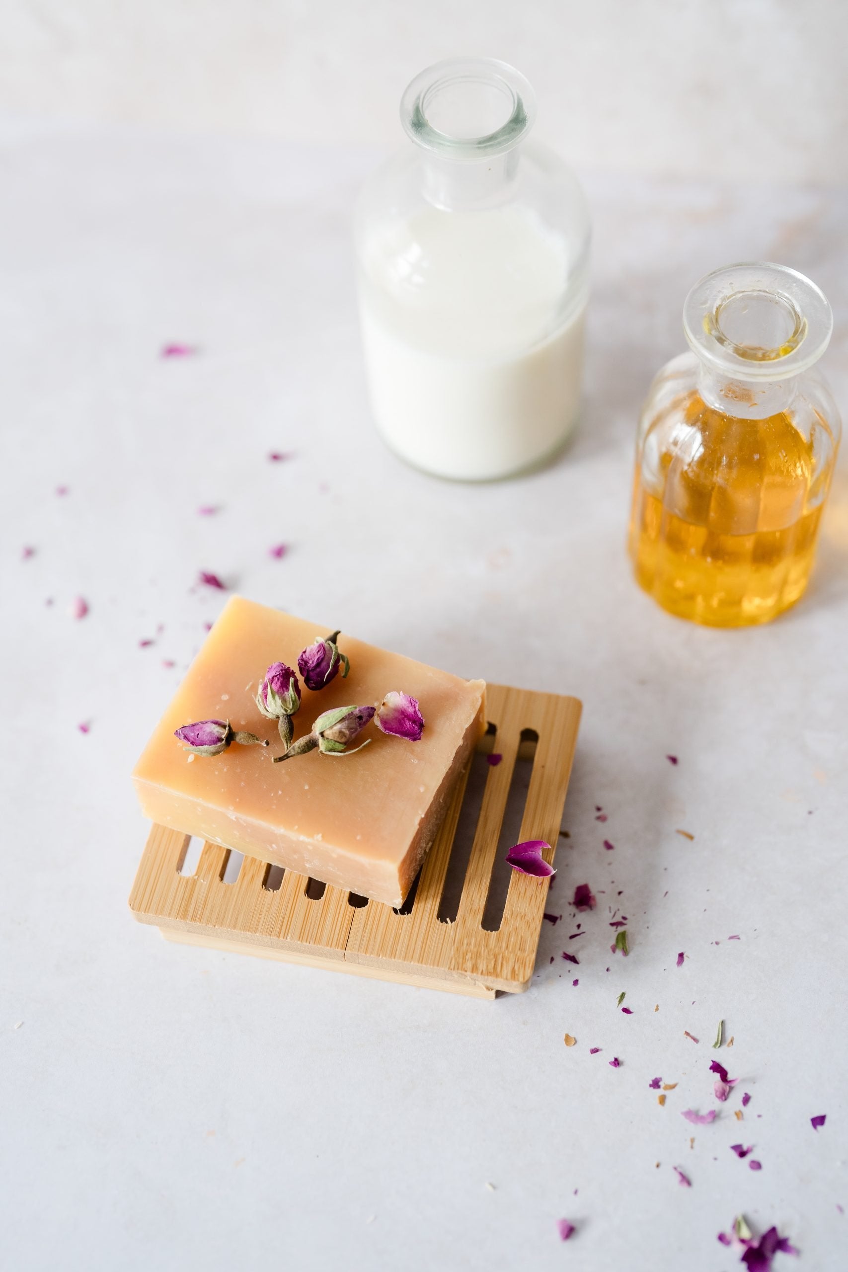 Bar of soap on a wooden soap dish with flowers, next to a bottle of milk and a bottle of oil on a light surface.