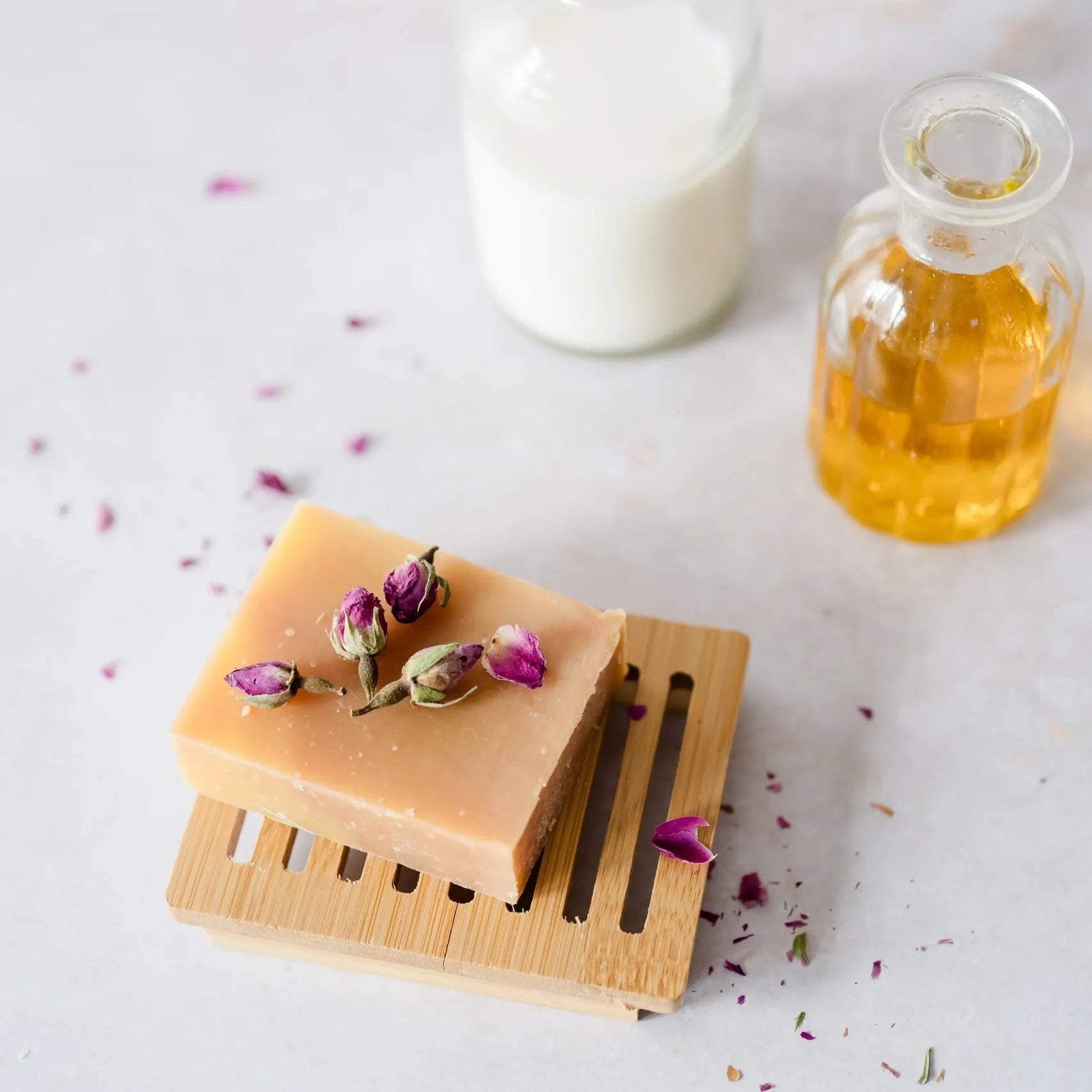 Bar of soap on a wooden soap dish with flowers, next to a bottle of milk and a bottle of oil on a light surface.